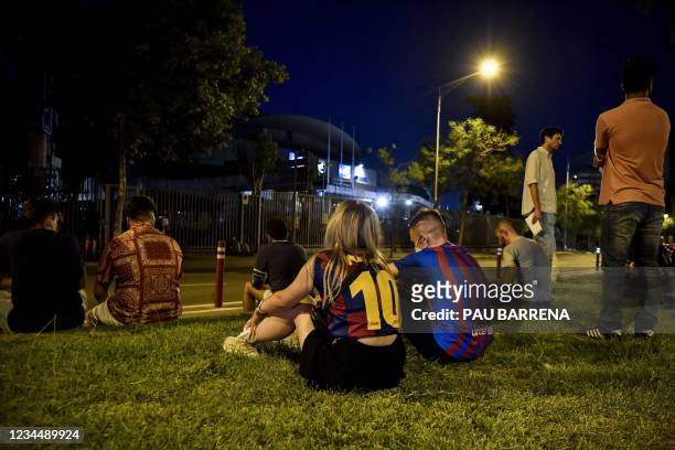Couple wearing jerseys of Barcelona's Argentinian forward Lionel Messi sit in the grass as supporters gather in front of the Camp Nou stadium in...