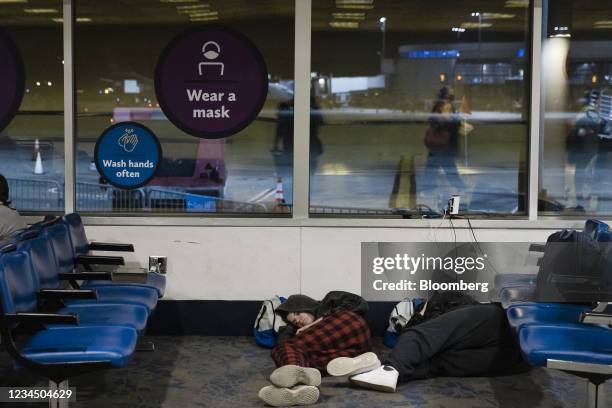 Travelers sleep under a "Wear A Mask" sign at Charlotte Douglas International Airport in Charlotte, North Carolina, U.S., on Thursday, Aug. 5, 2021....