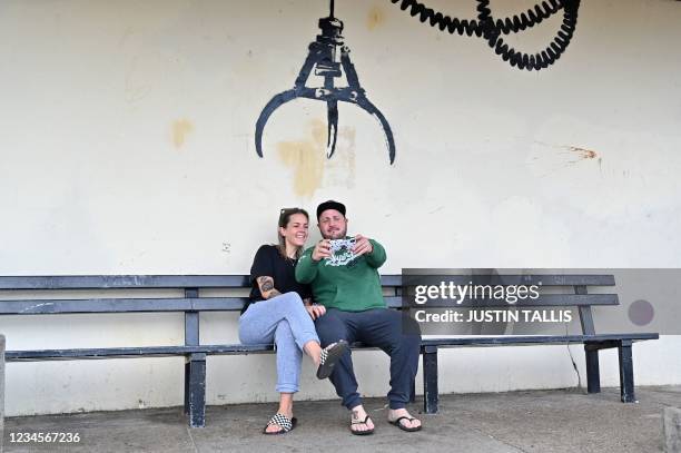 Couple take a selfie photograph below a graffiti artwork of an amusement arcade grabber, which bears the hallmarks of street artist Banksy, on a wall...