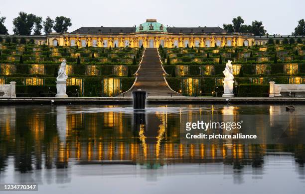 August 2021, Brandenburg, Potsdam: Sanssouci Palace is reflected in the morning in the water of the fountain on the main avenue at the foot of the...