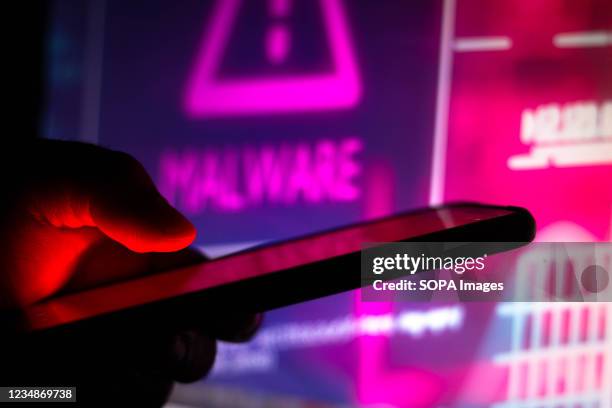 In this photo illustration the malware logo seen in the background of a silhouette hand holding a mobile phone.