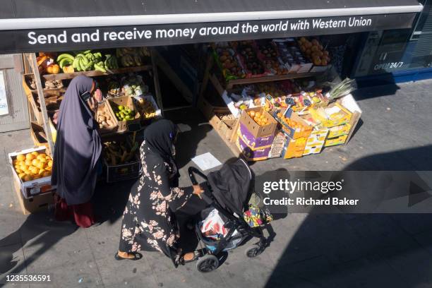 Two Musim women walk past a greengrocer selling fruit and veg and which is also advertising the transfer of money to and from abroad by Western...