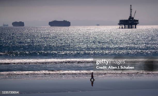 Huntington Beach, CA Container ships and an oil derrick line the horizon as a lone environmental oil spill cleanup crew member searches the beach,...