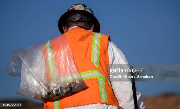 Huntington Beach, CA An environmental oil spill cleanup crew member wears a hot protective suit on a hot day as he walks the beach, loading chunks of...