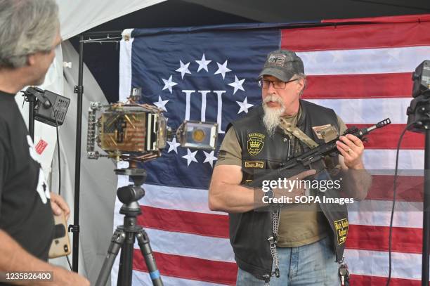 Man holding an automatic rifle poses in front of a 3% flag which is a far-right anti-government militia movement. The Rod of Iron Freedom Festival is...