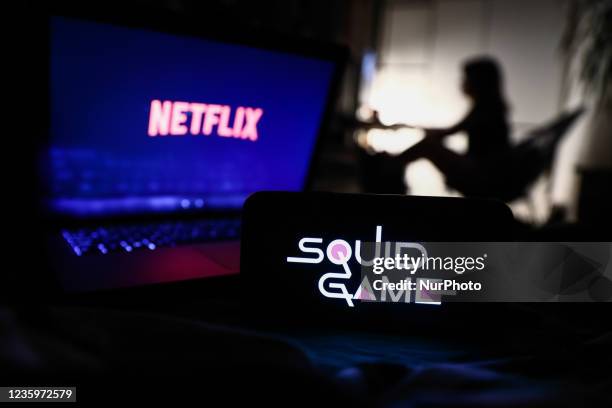 Squid Game series logo displayed on a phone screen, Netflix logo displayed on a laptop screen and a silhouette of a woman in the background are seen...