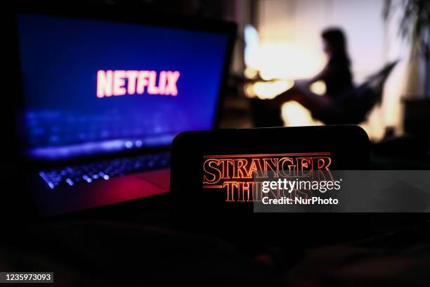 Stranger Things series logo displayed on a phone screen, Netflix logo displayed on a laptop screen and a silhouette of a woman in the background are...