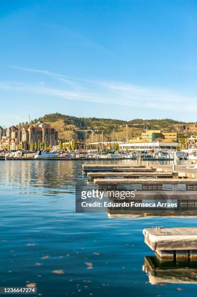 view of kelowna's downtown marina, boats, sailboats and mountains on the background, against a sunny clear blue sky - kelowna stock pictures, royalty-free photos & images