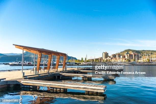 view of kelowna's downtown marina, boats, sailboats and mountains on the background, against a sunny clear blue sky - kelowna stock pictures, royalty-free photos & images