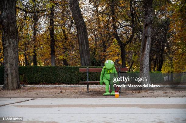 Man dressed as Kermit the Frog sits alone on a bench in the Retiro Park during an Autumn day, hoping to get some coins from visitors who take photos...