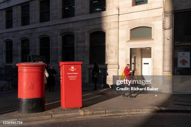 Woman wearing a red coat walks through winter sunlight and past two Royal Mail postboxes at the corner of King William and Lombards streets in the...