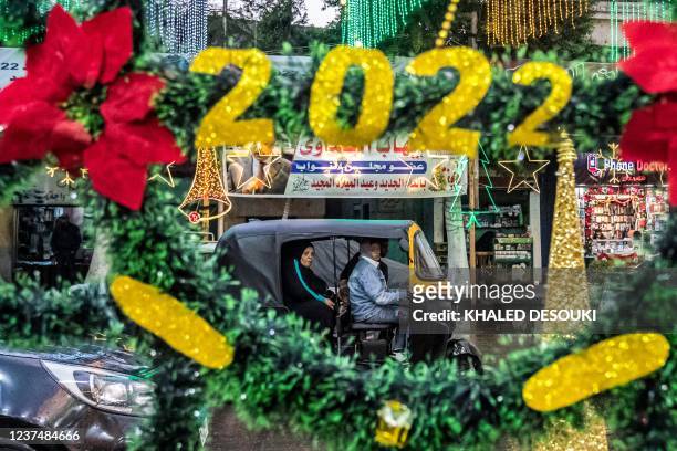 People ride in a tuk-tuk past Christmas decorations reading "2022" on display at a shop exhibiting Christmas accessories outside along a main street...