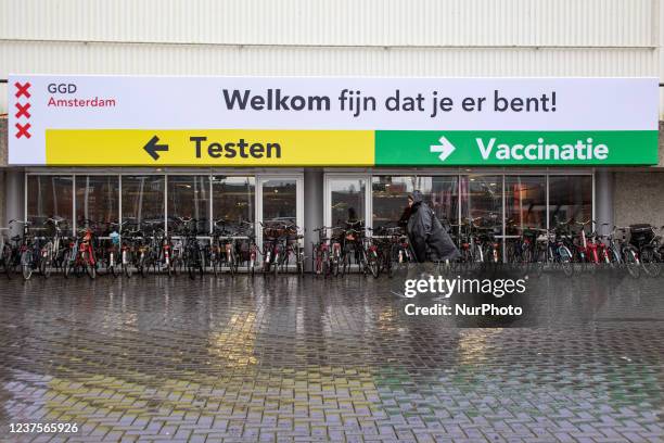 Woman as seen walking towards the testing facility under a banner with direction for testing or vaccination center. People wearing facemasks wait...