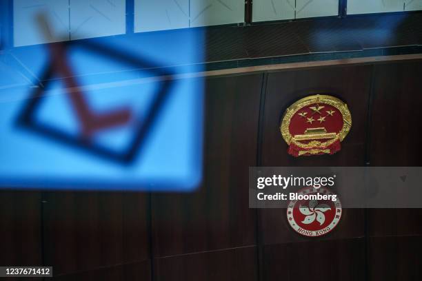 An image of voting tick reflected on a window as the newly-installed emblem of China, top, displayed above the emblem of the Hong Kong Special...