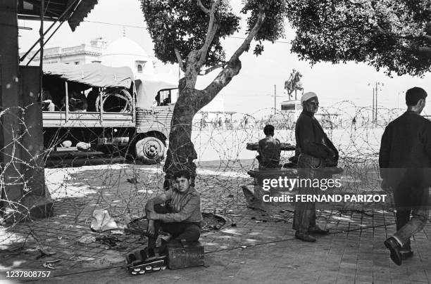 Young Algerian shoe shiner waits near barbed wire and a French army military truck in May 1962 on the Place du gouvernement in Algiers, while...