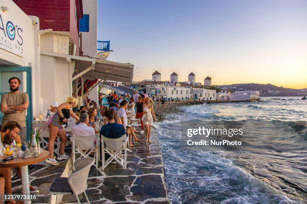 The iconic windmills of Mykonos island as seen from Little Venice with tourists at the bars, cafes and restaurants. Mykonos is an island in Greece in...
