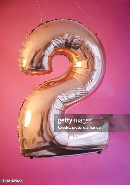 January 2022, Berlin: ILLUSTRATION - A golden balloon in the shape of the number 2 hangs in front of a pink background. The "2": symbol for...