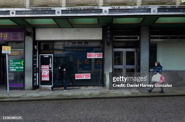 Hooded man holding a canned drink and a woman holding multiple bags of shopping walk past a shuttered high street shop with red and white closing...