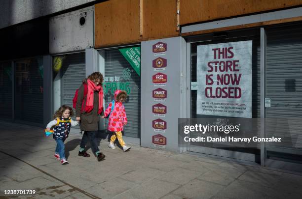 Woman and two children, all dressed in brightly coloured clothing, hold hands as they pass a permanently closed high street shop with a sign in the...