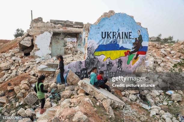 February 2022, Syria, Binnish: Syrian children sit next to a mural painted by painters in solidarity with Ukraine on a wall of a destroyed home in...