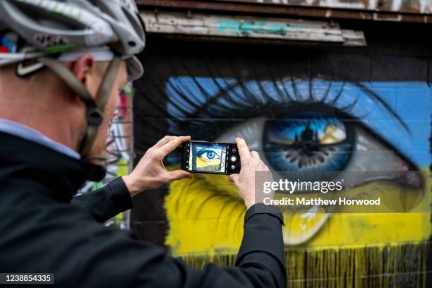 Man photographs graffiti artwork by MyDogSighs, from Southsea, Hampshire, showing a weeping eye in the colours of a Ukrainian flag in Northcote Lane...