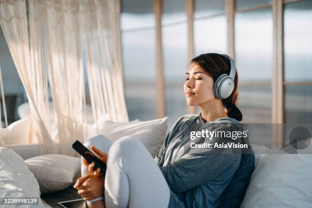 ontspannen jonge aziatische vrouw met gesloten ogenzitting op haar bed dat van muziek over hoofdtelefoons van smartphone thuis geniet - apartment living asian stockfoto's en -beelden