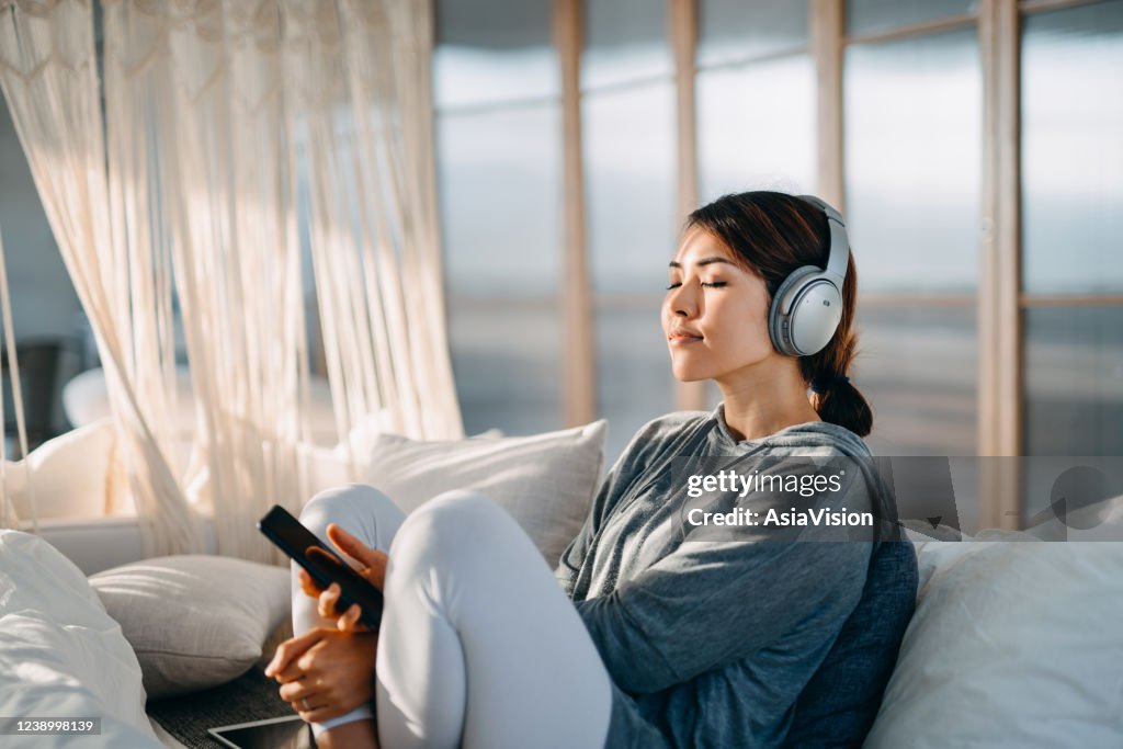 Relaxed young Asian woman with eyes closed sitting on her bed enjoying music over headphones from smartphone at home
