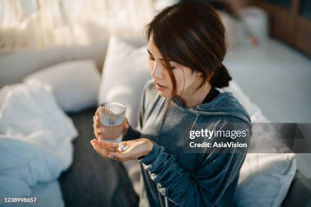 young asian woman sitting on bed and feeling sick, taking medicines in hand with a glass of water - diabetes pills imagens e fotografias de stock