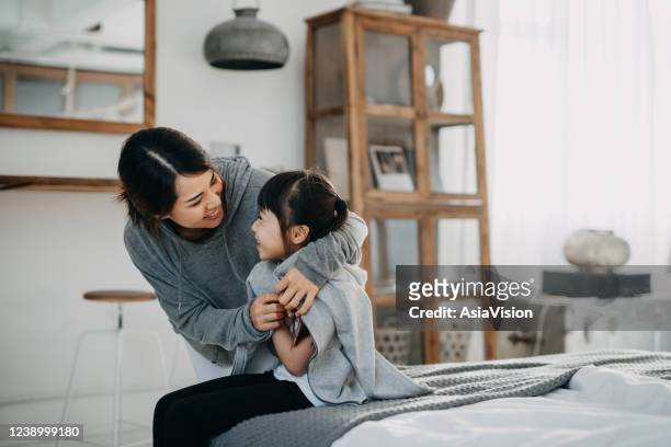 premurosa giovane madre asiatica che mette un cappotto su sua figlia a casa - freddo foto e immagini stock