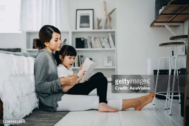 joven madre asiática sentada en el suelo en el dormitorio leyendo libro a la hija pequeña, disfrutando del tiempo de unión familiar en casa - familia en casa fotografías e imágenes de stock