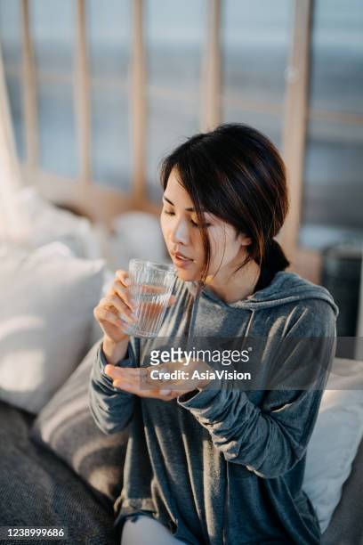 young asian woman sitting on bed and feeling sick, taking medicines in hand with a glass of water - diabetes pills stock pictures, royalty-free photos & images