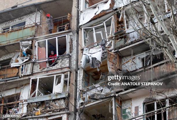 Resident uses a dustpan and broom to clear the debris from a flat, as another looks out of the destroyed front of a room, in a multi-storey building...