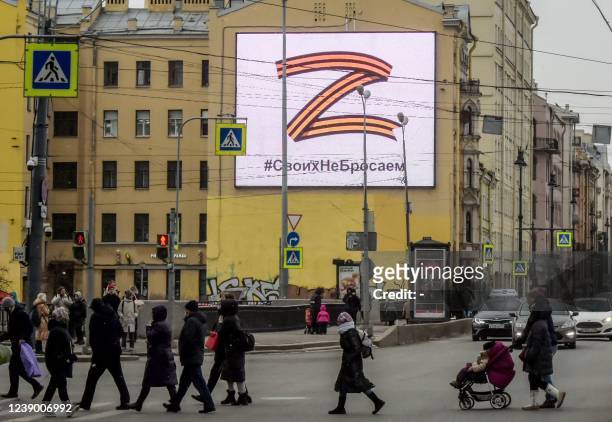 Pedestrians cross a street in front of a billboard displaying the symbol "Z" in the colours of the ribbon of Saint George and a slogan reading: "We...