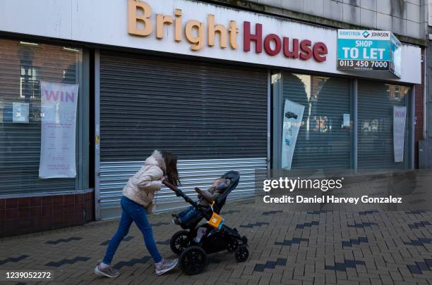 Young girl pushes a pram with a baby in it past a permanently closed branch of the former high street pay weekly chain BrightHouse that is now...