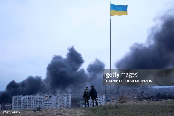 People standing near a Ukrainian national flag watch as dark smoke billows following an air strike in the western Ukrainian city of Lviv, on March...