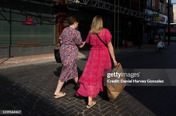 Two women, both wearing bright pink coloured floral dresses, and one carrying a brown paper carrier bag for fashion retail brand H&M, pass a closed...