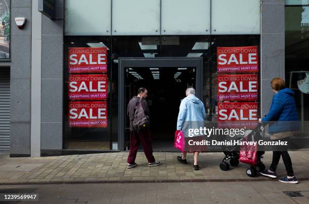 Man, an elderly woman, and a woman pushing a pram, the latter two carrying shopping bags, enter a shop advertising a closing down sale with window...