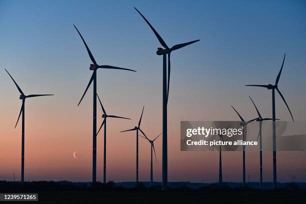March 2022, Brandenburg, Sieversdorf: Colorful glow of the morning sky and the crescent moon just before sunrise behind wind turbines in East...
