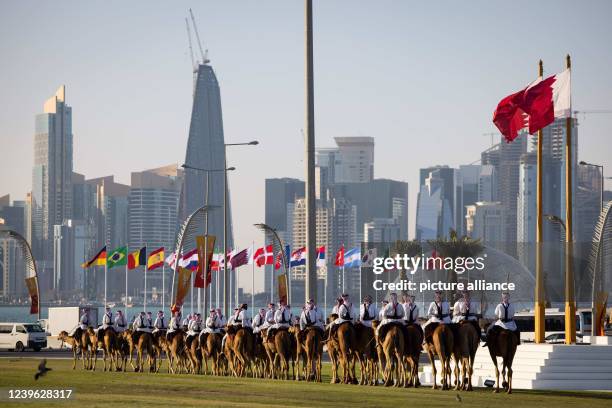 March 2022, Qatar, Doha: Riders on their camels are riding on the lawn in front of the parliament building on the Corniche promenade. The skyline of...