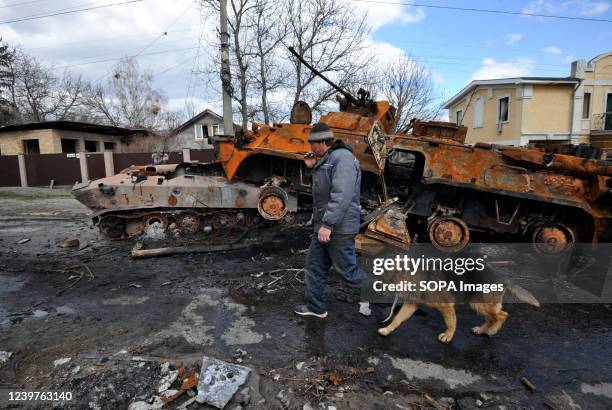 Man walks by with his dog past destroyed military equipment of the Russian army against the background of severe destruction in the city of Bucha....