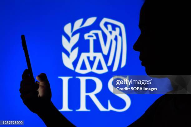 In this photo illustration, a woman's silhouette holds a smartphone with the Internal Revenue Service logo in the background.