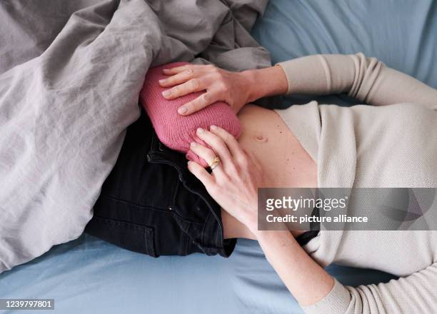 March 2022, Berlin: ILLUSTRATION - A woman holds a hot water bottle to her lower abdomen while lying in bed. Many sufferers of the abdominal disease...