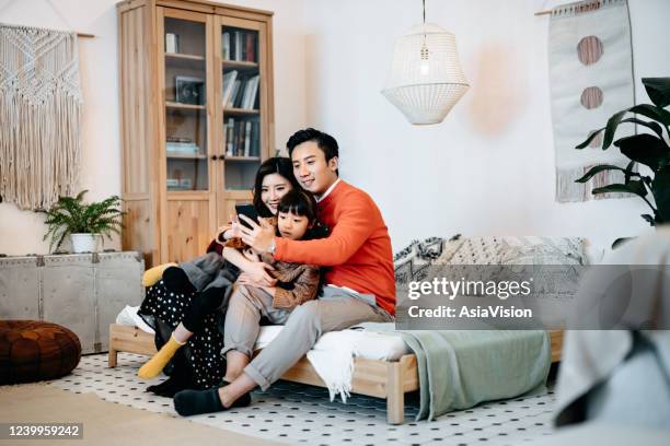 joyful young asian family using smartphone together. they are sitting on the bed in the bedroom video conferencing with friends and family - chinese family taking photo at home stock pictures, royalty-free photos & images