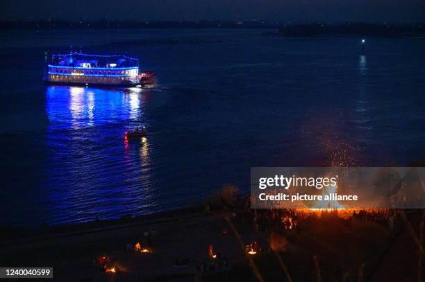 April 2022, Hamburg: The blue illuminated paddle steamer Louisiana Star sails along the Elbe behind one of the smaller Easter bonfires in Blankenese....