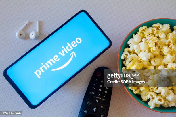 In this photo illustration, the Amazon Prime Video logo seen displayed on a smartphone along with a bowl of popcorn, headphones, and a tv remote.