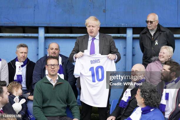 Prime Minister Boris Johnson holds a shirt with his name on next to the fans in the stands during a visit to Bury FC at their Gigg Lane ground, on...