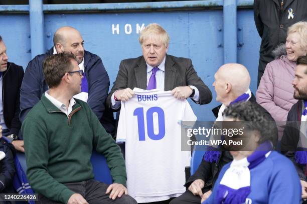 Prime Minister Boris Johnson holds a shirt with his name as he sits with fans during a visit to Bury FC at their Gigg Lane ground, on April 25, 2002...