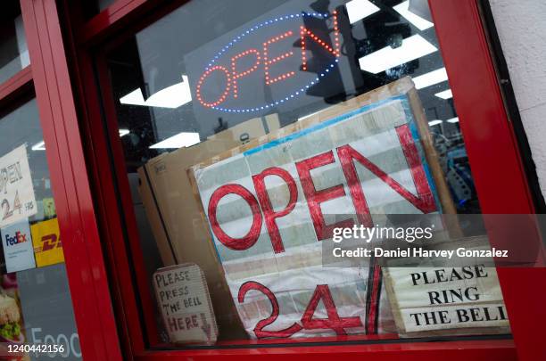 Numerous signs in a convenience shop window, one neon and others written by hand, advertise that the shop is open 24 hours a day, 7 days a week, on...