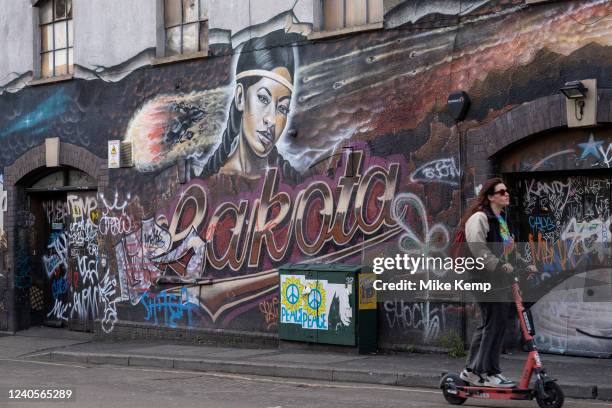 Woman on an eScooter passes the mural on the side of the famous Lakota club in Stokes Croft, one of the original homes of street art and graffiti, on...