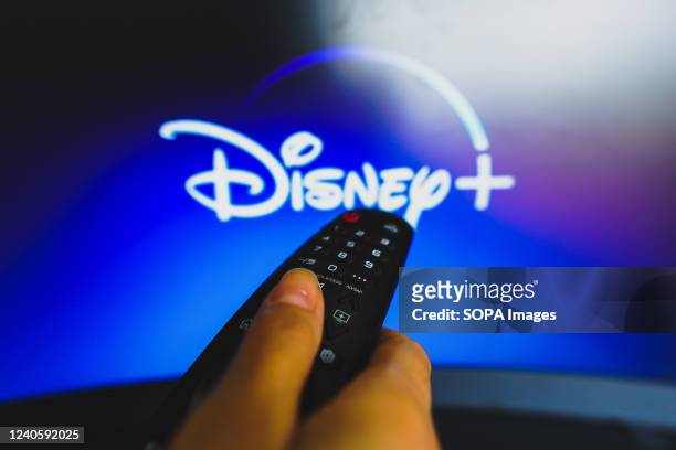 In this photo illustration, a hand holding a TV remote control in front of the Disney Plus logo on a TV screen.